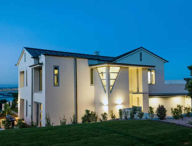 Exterior twilight view of a large contemporary two-storey residence with white rendered walls, dark standing-seam metal roofing, strong gabled roof forms, and a projecting entrance volume with a distinctive illuminated triangular window feature. The landscaped foreground includes lawn and low planting, while a body of water and distant shoreline are visible beyond the house.