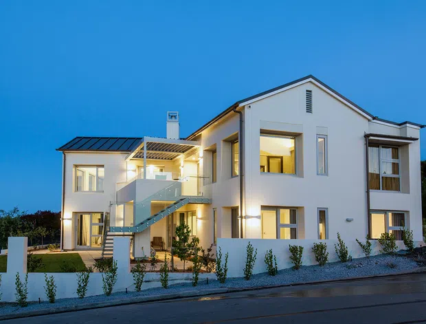 A contemporary multi-level white residential house photographed at dusk, featuring a gabled central volume, flat-roofed side wings, large windows, an external glazed stairway and balcony, warm interior lighting, and a low landscaped street frontage.
