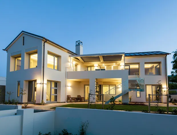 A large contemporary two-storey white residence photographed from the front at dusk. The home features a pitched roof over the left wing, a broad flat-roofed central outdoor living area, extensive glazing, recessed illuminated balconies and terraces, and an exposed external stair. A low white boundary wall and landscaped garden occupy the foreground.