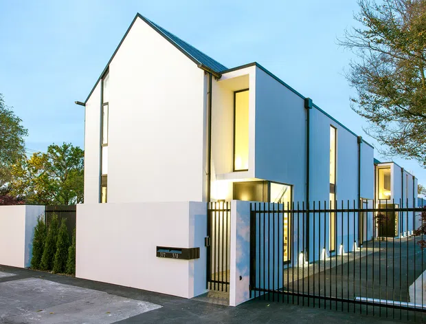 Exterior view of a contemporary multi-unit residential development with crisp white rendered walls, narrow vertical windows, projecting box forms, and steeply pitched dark metal roofs. A black metal security gate and fence enclose the paved entry and driveway, with warm interior lighting visible through the windows.