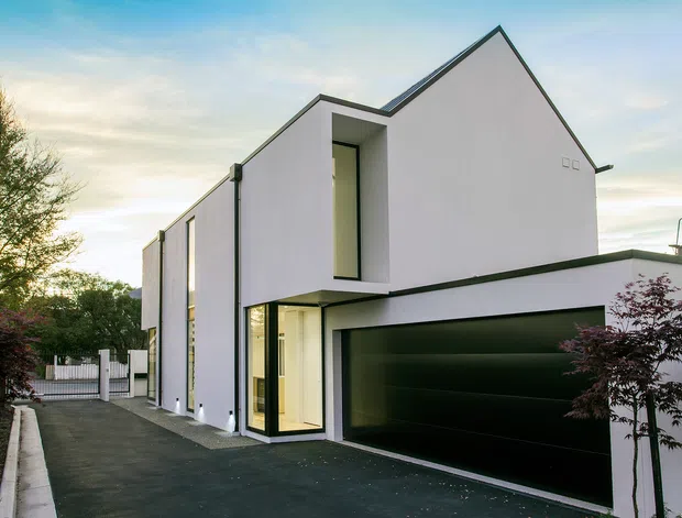 Contemporary white multi-level residential house viewed from the driveway, featuring sharp angular gable-like roof forms, clean geometric volumes, dark-framed glazing, and a large black integrated garage door. A narrow paved driveway and landscaped street frontage frame the facade.