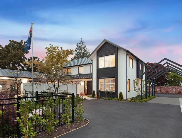 Exterior view of a contemporary multi-level residential building at dusk, with white rendered walls, dark charcoal cladding, pitched gabled roof forms, extensive windows, landscaped planting, a paved driveway, and a prominent open metal pergola or covered parking structure. A flagpole and fenced garden terrace are visible in the foreground.