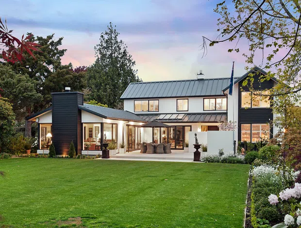 Exterior view of a large contemporary two-storey residence set within a landscaped garden. The home features dark vertical cladding, pale rendered walls, extensive glazing, a low-pitched metal roof, a covered patio, and a broad manicured lawn with established planting.