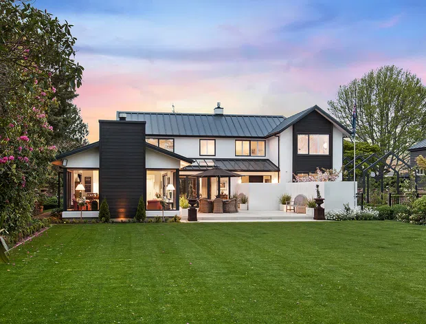 Exterior view of a large contemporary two-storey home set behind an expansive manicured lawn and landscaped garden. The house features dark vertical cladding, white rendered walls, a standing-seam metal pitched roof, large glazed openings, and a covered outdoor living area. Mature trees and planting frame the property beneath a pastel evening sky.