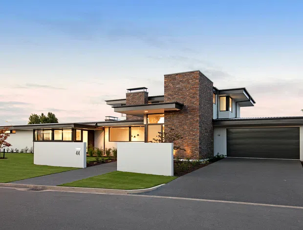 Contemporary two-storey residential home photographed from the street at dusk. The design features a strong vertical brick-clad volume, broad low-pitched roof planes, extensive dark-framed glazing, crisp white rendered walls, a landscaped front lawn, and an integrated double garage with a wide dark door.