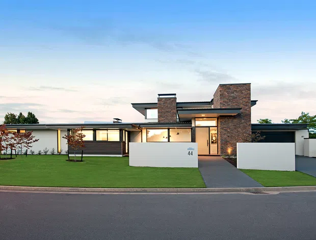 Street-facing exterior view of a contemporary single-storey residence with a low-pitched flat roof, dark-framed glazing, white rendered walls, and prominent brick-clad chimney volumes. The home has a manicured front lawn, paved entry path, boundary walls, and an integrated garage.