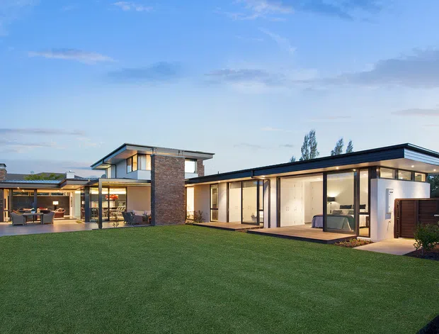 Exterior view of a contemporary single-storey residence with a low-pitched flat roofline, extensive floor-to-ceiling glazing, dark window frames, warm timber-lined soffits, and a stone-clad central volume. The home opens onto a broad manicured lawn and connected outdoor entertaining areas, photographed at dusk.