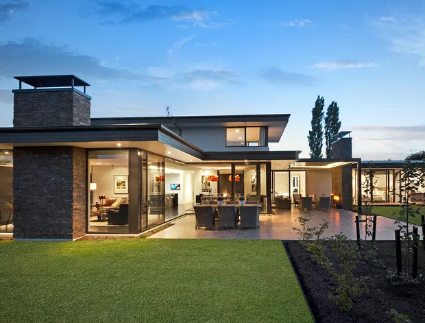 Exterior view of a contemporary, low-profile residence at dusk, with extensive floor-to-ceiling glazing, a projecting flat roof, dark horizontal cladding, and a prominent stone-clad chimney volume. The home opens onto a paved courtyard with outdoor seating and landscaped lawn and garden beds. Warm interior lighting and visible living spaces create an inviting connection between indoors and outdoors.
