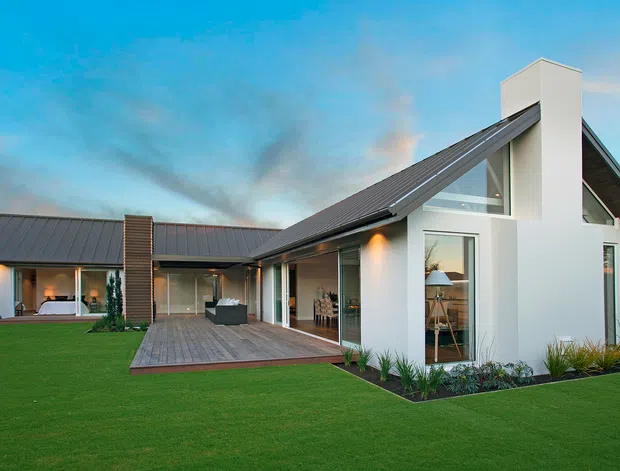 Exterior view of a contemporary single-storey home with a prominent pitched gable roof, white rendered walls, dark metal roofing, extensive glazing, and a tall chimney-like vertical feature. A timber deck connects the living spaces to a neatly landscaped lawn, with a covered outdoor entertaining area visible at the rear.