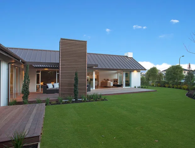 Exterior view of a low, modern single-storey home arranged around a broad landscaped lawn. The house has dark standing-seam metal pitched roofs, extensive floor-to-ceiling glazing, timber decking, a projecting vertical timber-clad feature wall, and sheltered outdoor living areas. The neat garden and crisp linear forms create a contemporary residential character.