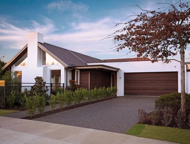 Contemporary single-storey suburban residence viewed from the street, featuring a steep gabled roof, white rendered walls, dark metal roofing, timber-look garage door and recessed entry. A low landscaped garden, paved driveway and street frontage frame the facade.