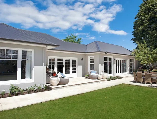 A bright, single-storey residential home viewed from a landscaped lawn and paved patio. The light-rendered exterior has a dark grey metal hipped roof, extensive white-framed multi-pane windows and French doors, and a broad paved terrace furnished with an outdoor dining setting and occasional seating. Formal garden beds and mature trees surround the home.