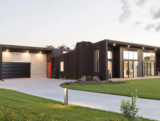 Contemporary single-storey residence with a low, angular flat-roofed form, dark vertical cladding, expansive glazed living areas, and an integrated garage. The home is viewed from a broad landscaped front lawn and concrete driveway in soft evening light.