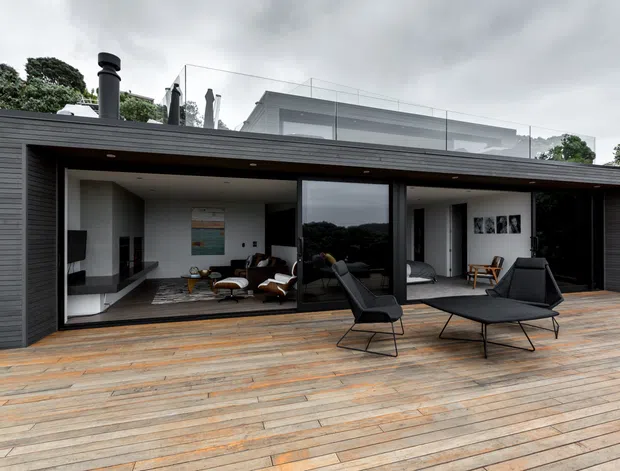 Contemporary single-storey pavilion-like home viewed from a large timber deck, with dark horizontal cladding, extensive floor-to-ceiling glazing, and a flat roof. The open interior reveals a living room with lounge seating, a media area, and a bedroom visible through the glazing. A second-level terrace or roof structure is visible behind the main volume, along with a chimney and glass balustrade. The scene is overcast and surrounded by mature greenery.
