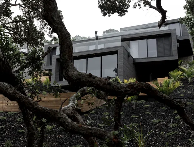 A contemporary multi-level house set into a landscaped, sloping site, viewed through mature tree branches. The dark grey exterior features horizontal cladding, large black-framed glazing, projecting box-like upper volumes, and a low-pitched or flat roofline. Timber retaining walls and native-style planting frame the approach.