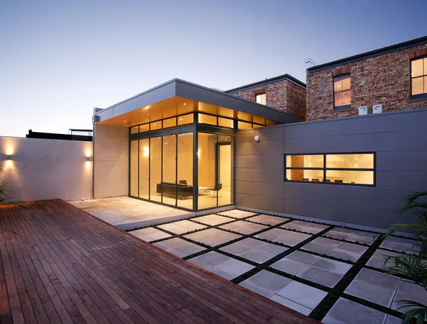 Twilight exterior view of a contemporary flat-roof building combining dark grey metal-clad volumes with an exposed red-brick upper structure. Floor-to-ceiling glazing forms a warmly illuminated corner pavilion, opening onto a timber deck and a geometric paved courtyard with dark grid joints. Minimal exterior lighting and tropical planting frame the modern outdoor space.