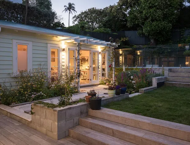 Exterior view of a renovated low-rise residential dwelling at dusk, featuring pale horizontal weatherboard cladding, illuminated glazed doors and windows, a covered pergola walkway, raised garden beds, timber decking, stepped timber seating, and landscaped planting. A dark privacy fence and garden structure sit behind the main building.
