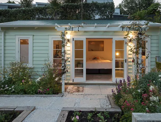 A compact single-storey garden dwelling with pale blue horizontal weatherboard cladding, a shallow corrugated metal roof and glazed French doors opening onto a paved path. Flowering garden beds and climbing plants frame the facade, while a bedroom with a bed is visible through the open doors. The setting has a calm, landscaped residential character.
