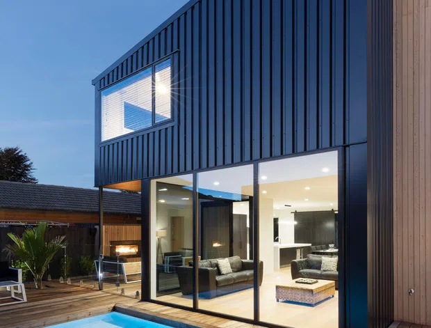 Dusk exterior view of a contemporary multi-level residential extension beside a brightly illuminated swimming pool. The building features a dramatic steep triangular roofline, dark vertical metal cladding, extensive floor-to-ceiling glazing, and a warm timber-clad side wall. A timber pool deck and outdoor seating connect the house to the pool, while the illuminated interior reveals a living area and kitchen.