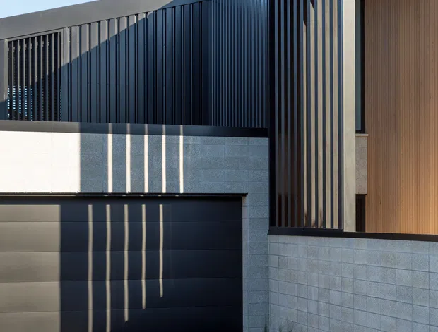 Contemporary residential facade viewed from the driveway, featuring a flat-roofed composition, dark vertical metal screening, pale masonry block walls, a wide dark garage door, and a warm timber-lined recessed entry or facade section. Strong sunlight creates linear shadows across the garage and walls.