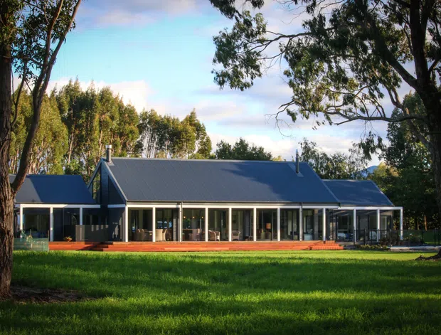 A single-storey modern residence set in a landscaped rural or suburban setting. The house has a long, low horizontal form, dark grey metal pitched roofs, extensive floor-to-ceiling glazing, and a broad timber deck or verandah along the front. Mature trees and open lawn frame the building, with a distant elevated landscape visible beyond.