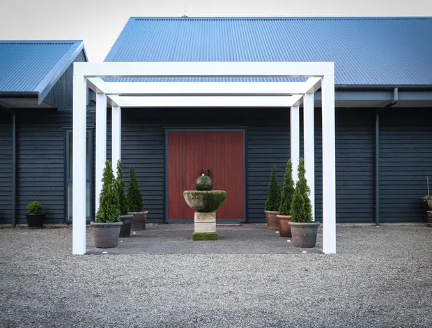 Front exterior view of a dark horizontal-clad building with multiple pitched metal roof volumes. A white rectangular entrance portico frames a tall red timber door, with symmetrical potted conifers and a central stone water feature on a broad gravel forecourt.