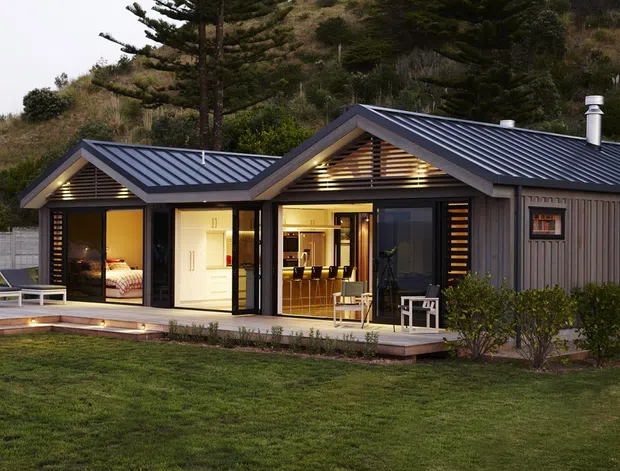 Single-storey contemporary coastal home set against a vegetated hillside, with two intersecting pitched gable roofs finished in dark metal, grey vertical cladding, extensive glazing, and a covered timber deck. Warm interior lighting reveals a bedroom and open-plan living or dining areas, while horizontal timber screening accents the gable ends.