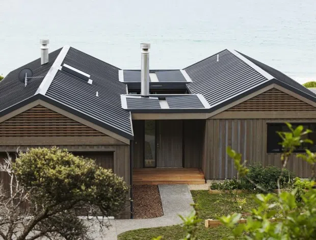 Exterior view of a low-profile coastal home with multiple dark pitched roof volumes, prominent metal flues, timber cladding, and a central recessed entry. A paved path crosses landscaped planting toward the house, with the ocean clearly visible behind it.