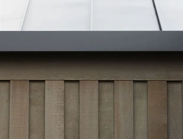Close-up architectural detail of a contemporary facade featuring vertical weathered timber battens beneath a dark horizontal metal fascia and a pale standing-seam metal upper panel. The composition emphasizes layered cladding, linear rhythm, and contrasting natural and industrial materials.