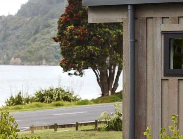 A close exterior view of a compact timber-clad building beside a landscaped roadside setting. The facade features vertical weathered timber boards, dark-framed windows, a simple metal gutter and downpipe, and a steeply pitched roof with broad dark eaves. A body of water and densely vegetated hills are visible beyond the road, giving the scene a distinctly coastal or lakeside character.