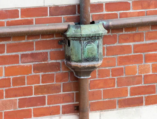 Close-up detail of a weathered, decorative green patinated metal rainwater or drainage junction mounted against a red brick wall. Horizontal and vertical brown metal pipes intersect at the ornate fitting, with light mortar joints and a pale rendered base visible at the bottom.