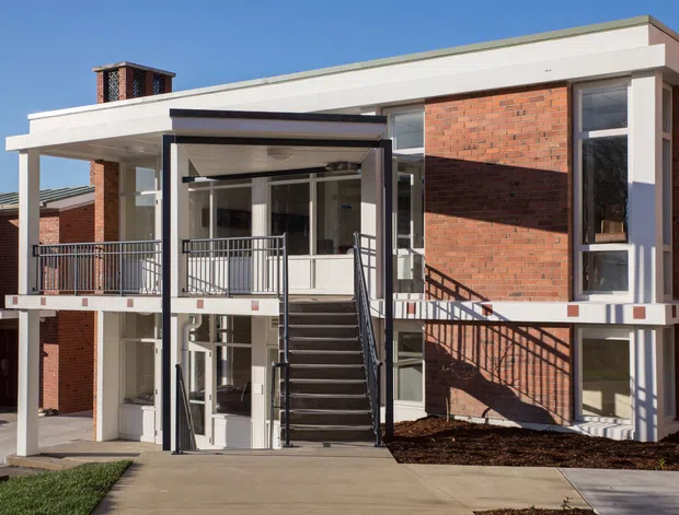 Exterior view of a contemporary two-storey brick building with a flat roof, white rendered structural framing, large glazed openings, an elevated balcony, and a central external stairway leading to the entrance. Black metal balustrades and a covered entry canopy provide contrasting details.