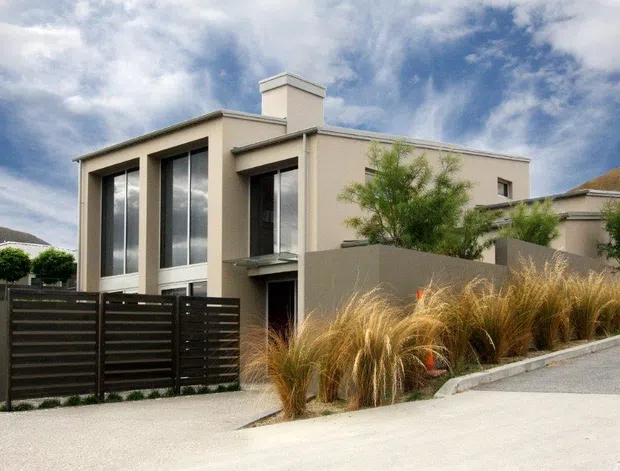Exterior view of a contemporary multi-storey home on a sloped site, with a clean rectilinear beige rendered facade, projecting box forms, tall dark-framed windows, a chimney-like rooftop element, and a dark horizontal-slat privacy fence. Ornamental grasses and small trees soften the boundary, while a paved driveway rises along the right side.