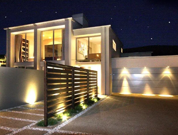 Night-time exterior view of a contemporary multi-level residence with strong rectilinear forms, large illuminated windows, a partially screened entry courtyard, horizontal timber-look privacy fencing, and an integrated double garage. Architectural lighting highlights the facade, driveway, and garage door.