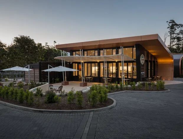A low-slung contemporary commercial building viewed from the landscaped forecourt at dusk. The building features a broad flat roof with deep overhangs, warm copper-toned curved cladding at the sides, extensive floor-to-ceiling glazing, a covered entrance terrace, outdoor umbrellas and seating, landscaped planting beds, and dark paved driveways.
