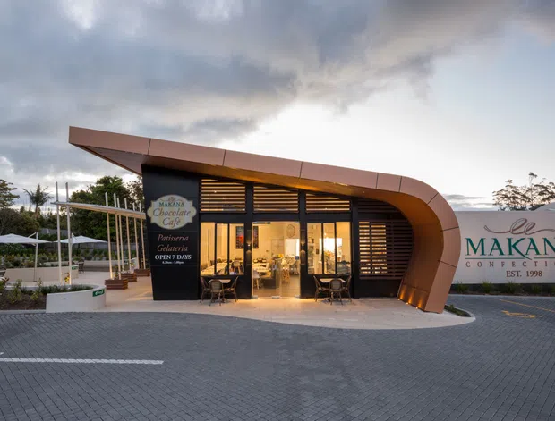 Exterior architectural photograph of the Makana Confections chocolate café and retail storefront at dusk. The low commercial building has a distinctive curved copper-toned canopy wrapping around a dark glazed entrance, horizontal timber or metal screening, warm interior lighting, and prominent branded signage. A paved forecourt and outdoor seating area are visible.
