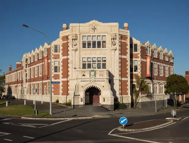 Street-level exterior photograph of a large historic institutional building on a prominent urban corner. The symmetrical façade combines red brick with pale stone or rendered detailing, ornate vertical brickwork, classical motifs, tall multi-pane windows, a stepped parapet roofline, and a grand arched entrance approached by stairs. Roads, traffic signage, street trees, and surrounding urban context are visible in the foreground.