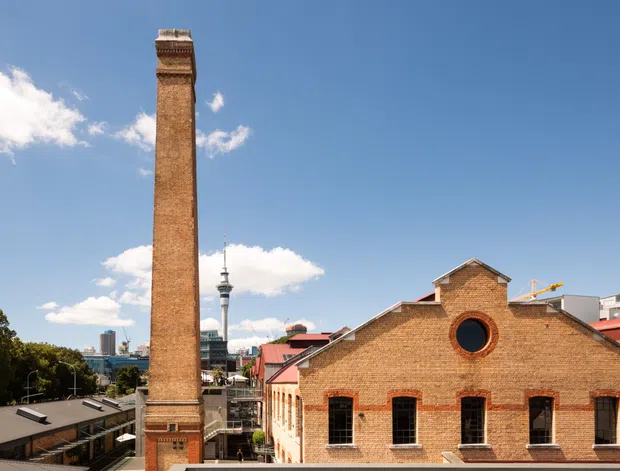 Exterior architectural view of a historic red-brick industrial building with a tall ornate chimney and gabled roofline. The facade includes arched and rectangular window openings, while a modern city skyline and distinctive tower are visible in the background under a bright blue sky.