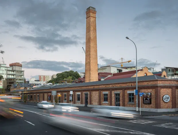 Urban exterior photograph of a historic red-brick industrial building with a tall brick chimney, arched brick detailing, dark metal roof, and rows of windows. The building sits on a city street with traffic light trails, modern buildings, and construction visible in the background.