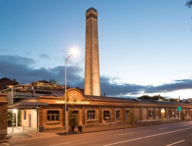 Twilight exterior view of a heritage-style brick commercial building on a wide urban street, dominated by a tall square brick chimney with a decorative cap. The low building has arched and rectangular window openings, detailed brickwork, warm interior lighting, and a rooftop terrace or landscaped outdoor area visible behind it. Streetlights, roadway markings, and surrounding urban context are visible.