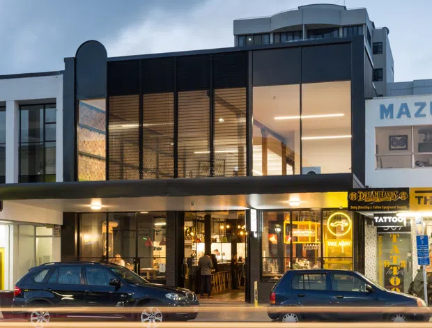 Street-facing view of a contemporary multi-storey commercial building at dusk. The main facade combines dark metal-framed glazing, black horizontal louvres, illuminated ground-floor retail or hospitality spaces, and a projecting upper-level volume with balconies. Adjacent storefront signage and parked cars line the urban street.