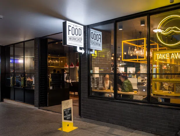 Street-level view of a compact contemporary food workshop or takeaway venue. The black-painted brick facade has large black-framed glazed windows and a glazed entry door, allowing views into the warmly lit commercial interior with counter service, bar shelving, pendant lights, signage, and prominent yellow neon graphics and takeaway lettering. The image is photographed from a covered pedestrian walkway.