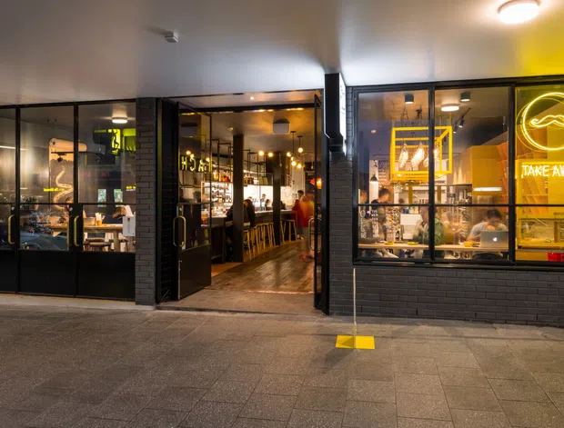 Street-facing view of a contemporary takeaway restaurant or café, with black brick cladding, extensive black-framed glazing, a warmly lit dining and service interior, bar seating, counter displays, and prominent yellow takeaway signage. The image is photographed from the exterior pavement, with the commercial storefront as the main subject.