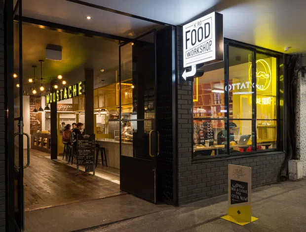 Night-time street-level view of a compact food workshop and takeaway venue, with a black brick facade, large glazed shopfront, illuminated signage, warm interior lighting, timber-look flooring, pendant lights, counter service area, and outdoor sidewalk signage. The image primarily presents the commercial building exterior while showing the food-service interior through the open entrance and windows.