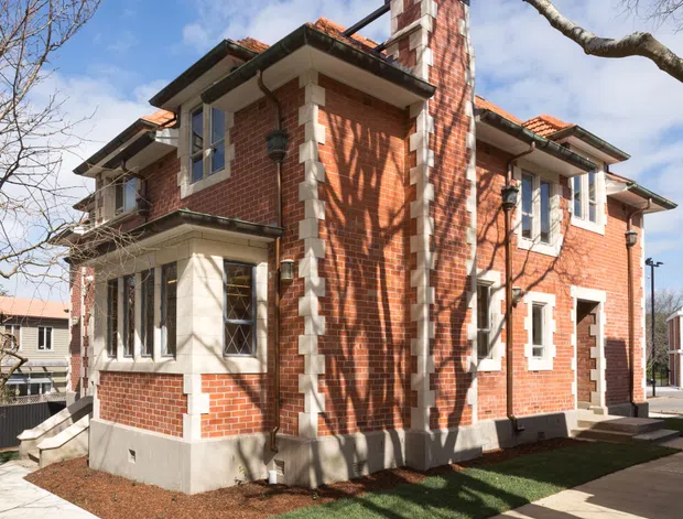 Exterior view of a compact multi-storey red-brick building with prominent white masonry quoins, trim and a tall chimney. The building has a tiled hipped roof, projecting bays, multi-pane windows, decorative brickwork and a small landscaped verge beside concrete footpaths. Leafless tree branches frame the sunny scene.