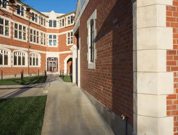 Exterior architectural photograph of a historic-looking red-brick institutional or commercial complex arranged around a landscaped courtyard. The buildings feature pale stone quoins and arched detailing, tall multi-pane windows, decorative masonry, and a paved pedestrian walkway under a clear blue sky.