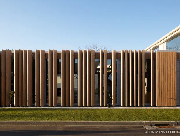 Contemporary single-storey building facade viewed from the street, featuring a rhythmic screen of closely spaced vertical timber fins over dark glazing and a timber-clad entrance door. A light-coloured roof edge and angular glazed upper section appear at the right, with a narrow lawn and pavement in front. The image is composed as an exterior architectural photograph in clear daylight.