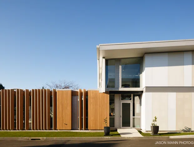 Street-facing exterior of a contemporary two-storey residence with a flat roof, crisp white and light-grey facade panels, extensive upper-level glazing, and a warm timber-slatted privacy fence and gate. A narrow lawn and concrete footpath sit between the house and street.