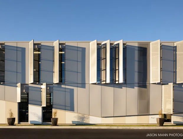 Street-facing exterior view of a contemporary multi-storey building with a highly regular facade of pale rendered or panelled surfaces, dark glazed openings, and projecting vertical fins or operable screens casting strong geometric shadows. The composition is minimalist and rectilinear, set against a clear blue sky.