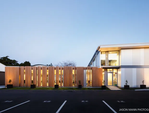 Exterior architectural photograph of a contemporary low-rise building viewed from a paved parking area at dusk. The facade combines smooth light-coloured panels with dark horizontal bands, extensive glazing, and a tall vertical timber-slat privacy or entry screen with warm interior lighting behind it. A flat roofline, clean geometric massing, planters, and marked parking bays are visible.